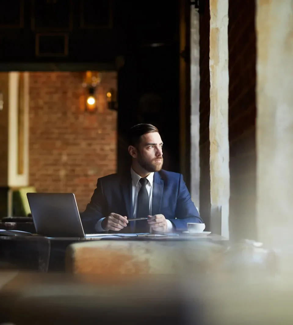 A man in a suit pausing over his laptop in a quiet loft restaurant, looking toward the window