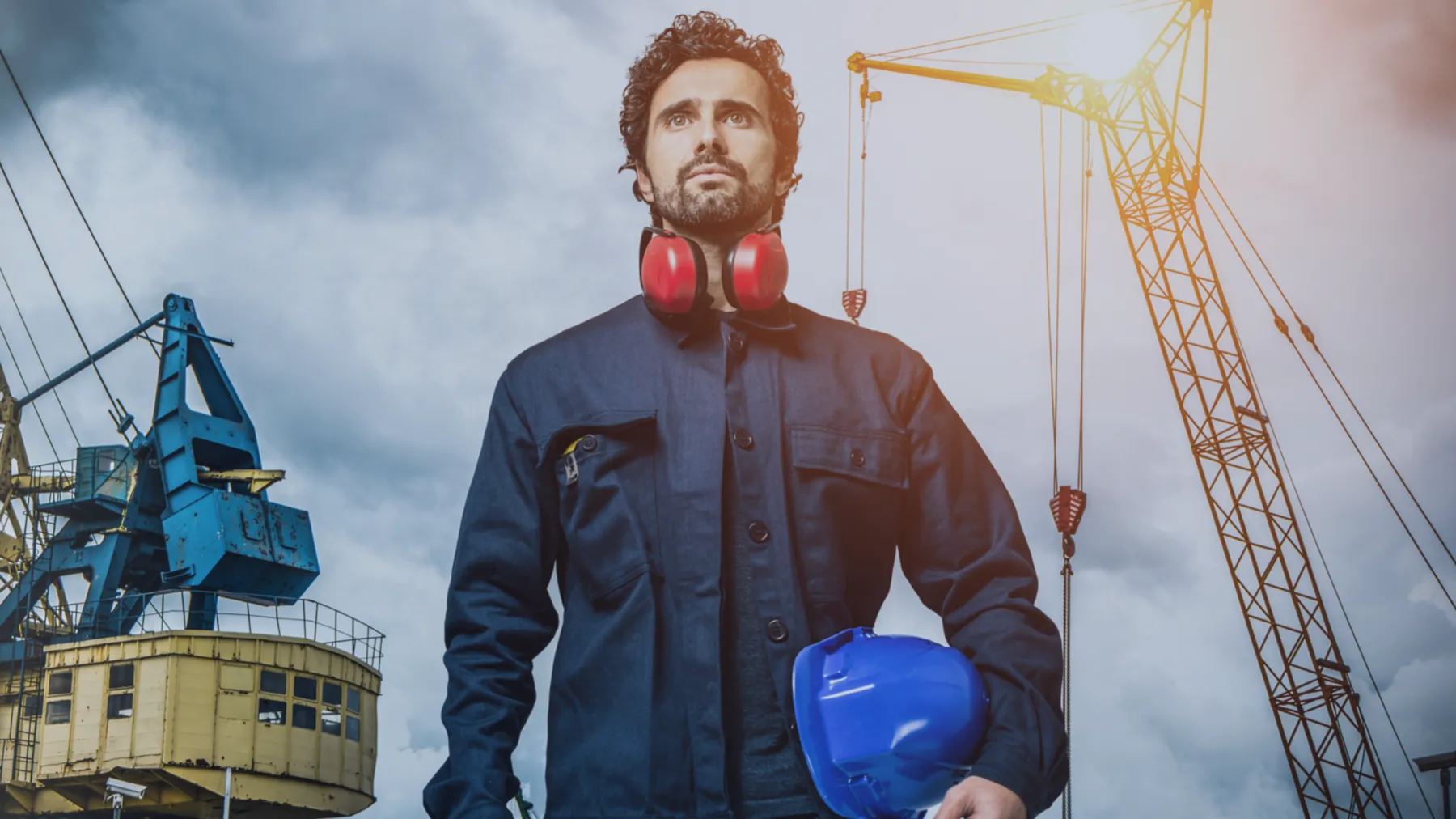A construction worker holding a hard hat before cranes